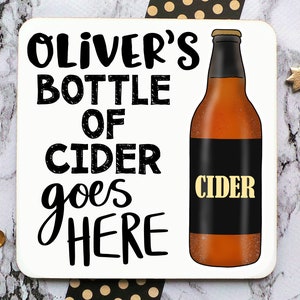 May include: A white coaster with black text that reads "Oliver's Bottle of Cider goes here". A brown bottle of cider with a black label that reads "Cider" is pictured on the right side of the coaster.