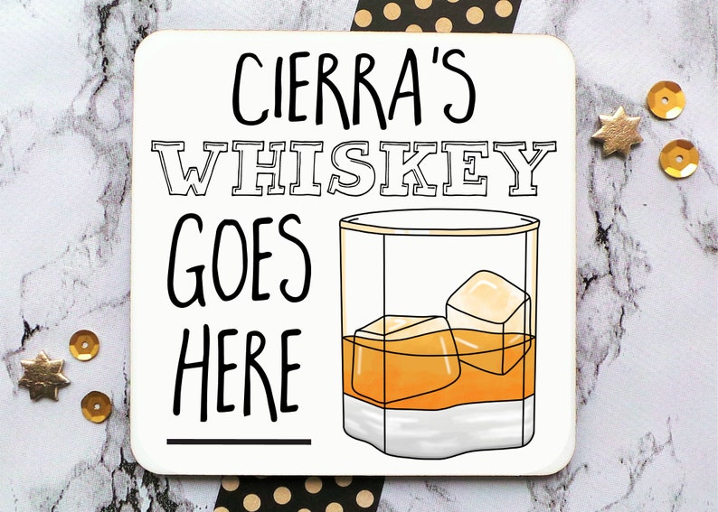 May include: White coaster with black text that reads "Cierra's Whiskey Goes Here" with a line underneath the text. A cartoon drawing of a glass of whiskey with ice cubes is to the right of the text.