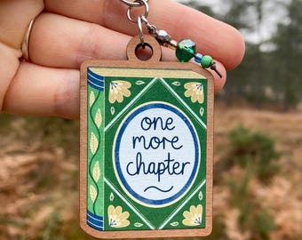 May include: A wooden book-shaped keychain with a green cover and gold accents. The text "one more chapter" is written in black on the front of the keychain.