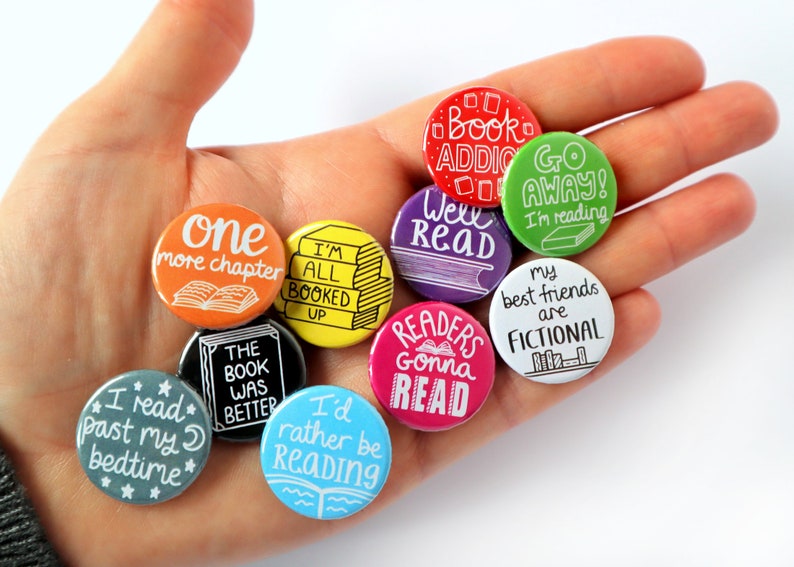 May include: A collection of ten round, colorful button pins with book-themed sayings. The pins feature phrases like "One more chapter", "I'm all booked up", "Well Read", "Readers gonna read", and "My best friends are fictional".