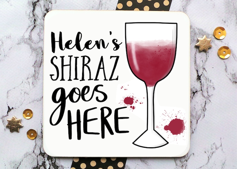 Puede incluir: Posavasos blanco con texto negro que dice "Helen's Shiraz goes HERE" y un dibujo de contorno negro de una copa de vino con vino tinto dentro. Hay salpicaduras de vino tinto en el posavasos.