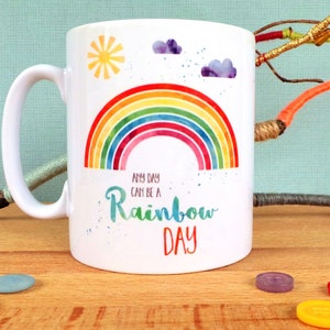 Puede incluir: Taza de cer&aacute;mica blanca con un dise&ntilde;o de arco&iacute;ris y el texto "Any day can be a rainbow day".