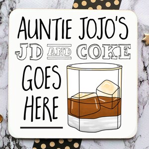 May include: A white coaster with black text that reads "Auntie Jojo's JD and Coke goes here" with a line underneath the text. A cartoon drawing of a glass of brown liquid with ice cubes is to the right of the text.