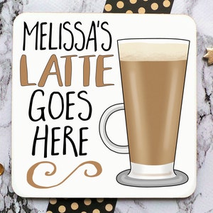 Op de afbeelding: Witte onderzetter met zwarte tekst die "Melissa's Latte Goes Here" luidt en een bruine illustratie van een latte in een glas. De onderzetter heeft een bruin swirl-ontwerp in de rechterbenedenhoek.