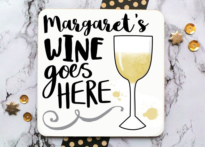 Puede incluir: Posavasos blanco con texto negro que dice "Margaret's Wine goes HERE" y un contorno negro de una copa de vino blanco. La copa est&aacute; llena de un l&iacute;quido amarillo claro.