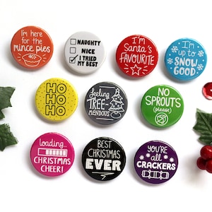 May include: Ten round Christmas themed buttons with various sayings. The buttons are in different colors, including red, green, blue, yellow, pink, black, and gray. The sayings include "I'm here for the mince pies", "I'm Santa's Favorite", "I'm up to snow good", "Ho Ho Ho", "Feeling Tree-mendous", "No Sprouts (please)", "Loading... Christmas Cheer", "Best Christmas Ever", and "You're all Crackers".
