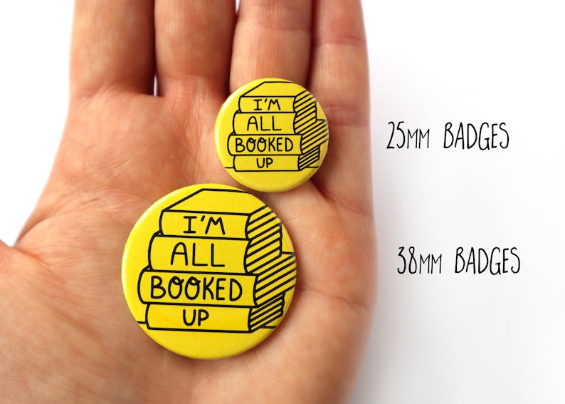 May include: Two yellow round badges with black outlines. The larger badge is 38mm and the smaller badge is 25mm. Both badges feature a stack of books with the text "I'm All Booked Up".