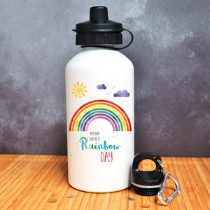 Puede incluir: Botella de agua de aluminio blanca con un arco&iacute;ris, nubes y un sol. El texto "Any day can be a Rainbow Day" est&aacute; impreso en la botella.