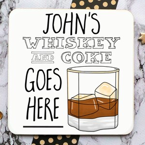 May include: White coaster with black text that reads "JOHN'S WHISKEY AND COKE GOES HERE" with a black and white illustration of a glass of whiskey with ice cubes.