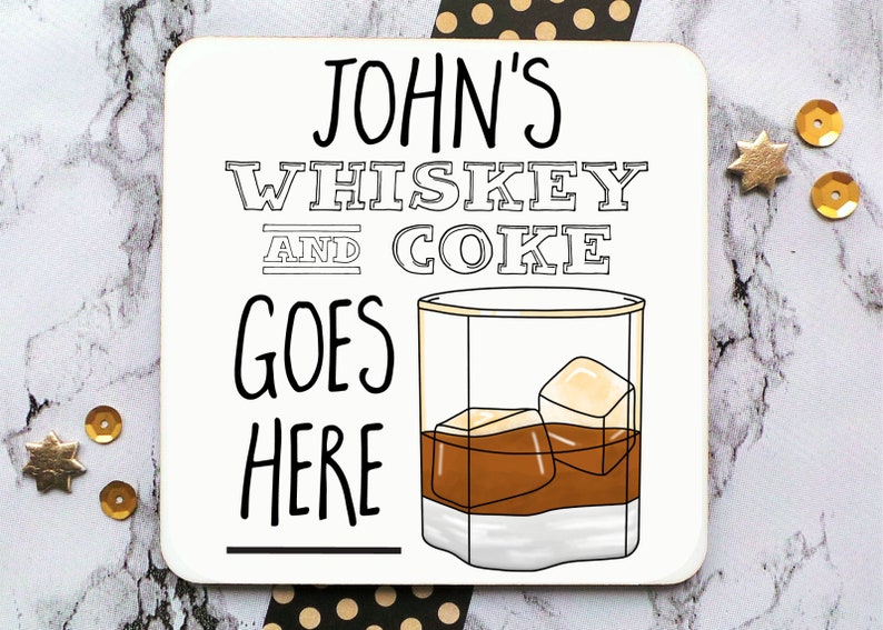 May include: White coaster with black text that reads "JOHN'S WHISKEY AND COKE GOES HERE" with a black and white illustration of a glass of whiskey with ice cubes.
