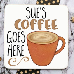 Op de afbeelding: Witte onderzetter met bruine tekst die "Sue's Coffee Goes Here" luidt en een bruine cartoon koffiemok met een swirl van room erbovenop.