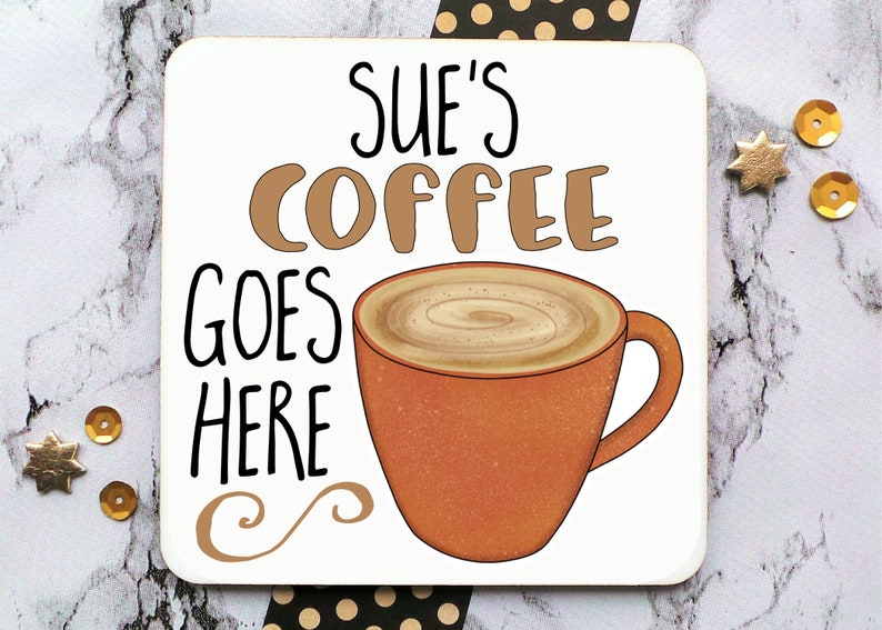 Op de afbeelding: Witte onderzetter met bruine tekst die "Sue's Coffee Goes Here" luidt en een bruine cartoon koffiemok met een swirl van room erbovenop.
