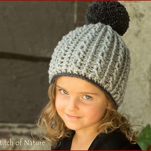 Puede incluir: Un gorro de punto gris y negro con un pompón en la parte superior. El gorro tiene un patrón de punto de cable y un ribete gris. El gorro lo lleva puesto un niño con el pelo largo y rizado.