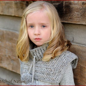 May include: A young girl with long blonde hair wearing a gray crocheted cowl with a pom-pom detail. The cowl has a tie at the neck. The girl is standing in front of a wooden wall.