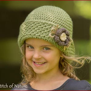 Puede incluir: Un sombrero cloche de ganchillo verde claro con una flor decorativa. El sombrero está adornado con una flor marrón y crema. El sombrero está hecho de hilo texturizado y tiene un ala. El texto "Stitch of Nature" está en la esquina inferior izquierda.