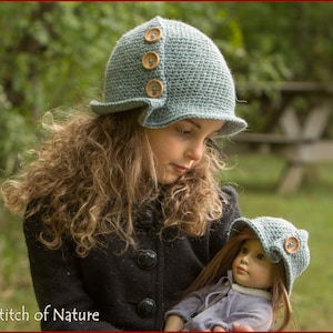 May include: A young girl wearing a blue crocheted hat with wooden buttons holds a doll wearing a similar hat. Both hats have a wide brim and a slightly floppy shape. The girl has long, curly brown hair and is wearing a black jacket.