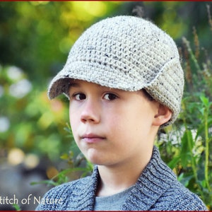 May include: A young person wearing a gray crocheted hat with a brim and a gray sweater. The hat has a unique design with a flap on the side.