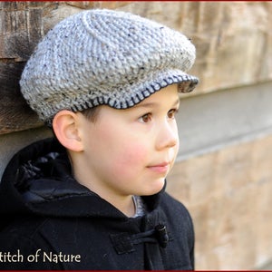 May include: A young person wearing a gray and black newsboy cap with a black trim. The cap is made of a soft, textured fabric. The person is wearing a black jacket with a hood.