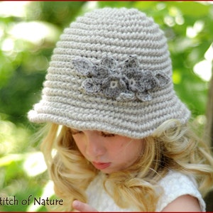 May include: A light gray crocheted hat with a small brim and a decorative gray floral embellishment. The hat is handmade and appears to be made of soft yarn. The hat is shown on a person. The text "Stitch of Nature" is in the bottom left corner.