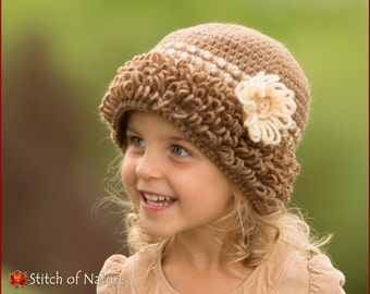 May include: A young girl wearing a brown crocheted hat with a white flower accent. The hat has a ruffled brim and a white stripe.