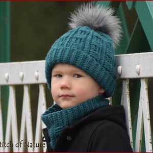 Puede incluir: Un gorro de punto azul turquesa con un pompón gris y una bufanda a juego. El gorro y la bufanda los lleva puestos un niño de pie delante de una valla metálica blanca.