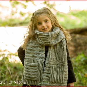 May include: A young girl wearing a gray crocheted scarf with pockets. The scarf is wrapped around her neck and tied in a loose knot. She is standing in a forest setting with green foliage and trees in the background.