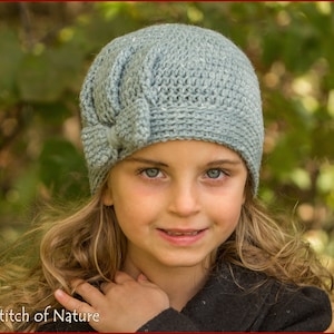 May include: A young girl wearing a light blue crocheted hat with a bow on top. The hat has a textured stitch pattern. The girl is smiling and looking at the camera.