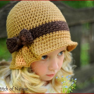 Puede incluir: Una niña pequeña lleva un gorro de ganchillo marrón y beige con un lazo marrón. El gorro tiene un ala ancha y está doblado hacia abajo en un lado. La niña está mirando hacia abajo a un ramo de flores azules.