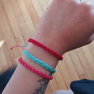 Puede incluir: Se muestran tres pulseras trenzadas en rojo, turquesa y rosa. Las pulseras están hechas de un material tejido y se llevan en la muñeca. Las pulseras son sencillas y coloridas.