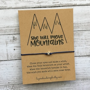 May include: A brown card with a blue string bracelet and a silver star charm. The card has a drawing of mountains and the text "She Will Move Mountains". The card also includes instructions for making a wish with the bracelet: "Close your eyes and make a wish, then tie this bracelet on your wrist. When the bracelet breaks in two, the wish you made will soon come true."
