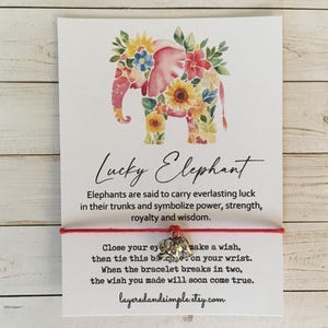 Pode incluir: Cartão com uma ilustração em aquarela de um elefante rosa adornado com flores, o texto "Lucky Elephant" e uma pulseira vermelha com um pequeno pingente de elefante. O texto explica a sorte e os desejos.