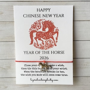 May include: A white card with red text and a red horse illustration, celebrating the Chinese New Year and the Year of the Horse 2026. A red string with a horse charm is attached. The text reads: "Close your eyes, then tie this bracelet on your wrist. When the bracelet breaks in two, the wish you made will soon come true."