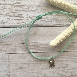 Pode incluir: Uma pulseira de cordão em verde menta claro com um pequeno pingente de metal prateado em forma de tartaruga marinha. A pulseira inclui um mecanismo de fecho de nó deslizante ajustável, permitindo um ajuste flexível no pulso ou no tornozelo.