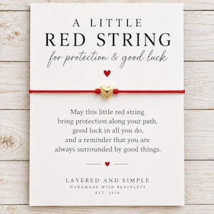 May include: A thin red string bracelet featuring a small, polished gold-tone heart charm, mounted on a white display card with black text that reads A LITTLE RED STRING for protection & good luck and includes a printed message and brand name.