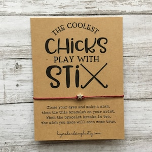 Könnte beinhalten: Rotes Armband mit einem silbernen Stern-Anhänger und einer Karte mit der Aufschrift "The Coolest Chicks Play With Stix". Die Karte enthält auch Anweisungen, wie man einen Wunsch an das Armband macht.