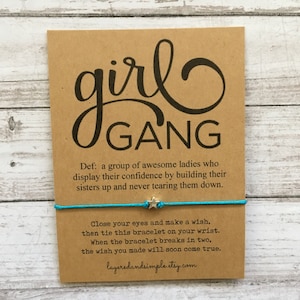 Op de afbeelding: Een bruine kaart met de woorden "girl GANG" in zwart schrift. Een turquoise armband met een sterbedel is bevestigd. De kaart bevat een definitie en instructies.
