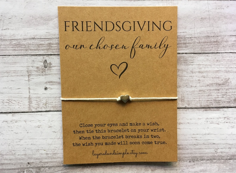 May include: A brown card with the text "FRIENDSGIVING our chosen family" and a heart symbol. A white string with a silver heart charm is tied around the card. The text "Close your eyes and make a wish, then tie this bracelet on your wrist. When the bracelet breaks in two, the wish you made will soon come true." is printed on the card. The text "layeredandsimpleetsy.com" is printed at the bottom of the card.