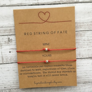 K&ouml;nnte beinhalten: Zwei rote Armb&auml;nder mit silbernen Perlen, beschriftet mit "Mine" und "Yours". Die Karte tr&auml;gt die Aufschrift "Red String of Fate" und enth&auml;lt ein Zitat &uuml;ber einen unsichtbaren roten Faden, der diejenigen verbindet, die dazu bestimmt sind, sich zu treffen.