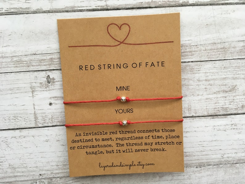 K&ouml;nnte beinhalten: Zwei rote Armb&auml;nder mit silbernen Perlen, beschriftet mit "Mine" und "Yours". Die Karte tr&auml;gt die Aufschrift "Red String of Fate" und enth&auml;lt ein Zitat &uuml;ber einen unsichtbaren roten Faden, der diejenigen verbindet, die dazu bestimmt sind, sich zu treffen.