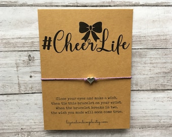 May include: A brown card with the text "#Cheer Life" and a black bow on top. A purple string bracelet with a silver heart charm is attached to the card. The card also includes instructions for making a wish and tying the bracelet on your wrist.