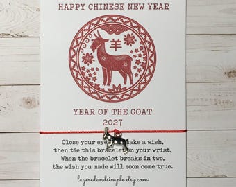 May include: A red string bracelet with a silver-toned goat charm attached to a white card. The card features a red circular graphic of a goat, the text HAPPY CHINESE NEW YEAR, YEAR OF THE GOAT 2027, and instructions for a wish-granting bracelet.