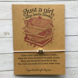 以下が含まれることがあります： 「Just a girl who loves books」という文字が書かれた茶色のカードと、ハートが描かれた本の積み重ね。カードには、金色のチャームが付いた白い紐が付いています。画像の下のテキストには、「目を閉じて願い事をし、このブレスレットを手首に結びます。ブレスレットが二つに割れたら、願い事が叶います。」と書いてあります。