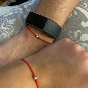 K&ouml;nnte beinhalten: Zwei rote Armb&auml;nder mit silbernen Perlen an jedem Handgelenk. Das Armband am linken Handgelenk wird mit einem schwarzen Fitness-Tracker getragen.