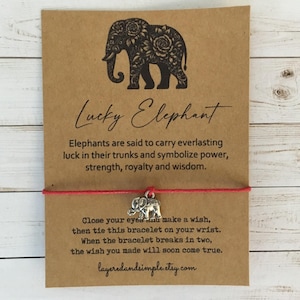 Pode incluir: Uma pulseira de desejos Lucky Elephant em um cartão marrom. O cartão apresenta uma ilustração de elefante preto e texto sobre elefantes que simbolizam sorte, poder e sabedoria. A pulseira tem uma corda vermelha e um pequeno pingente de elefante prateado.