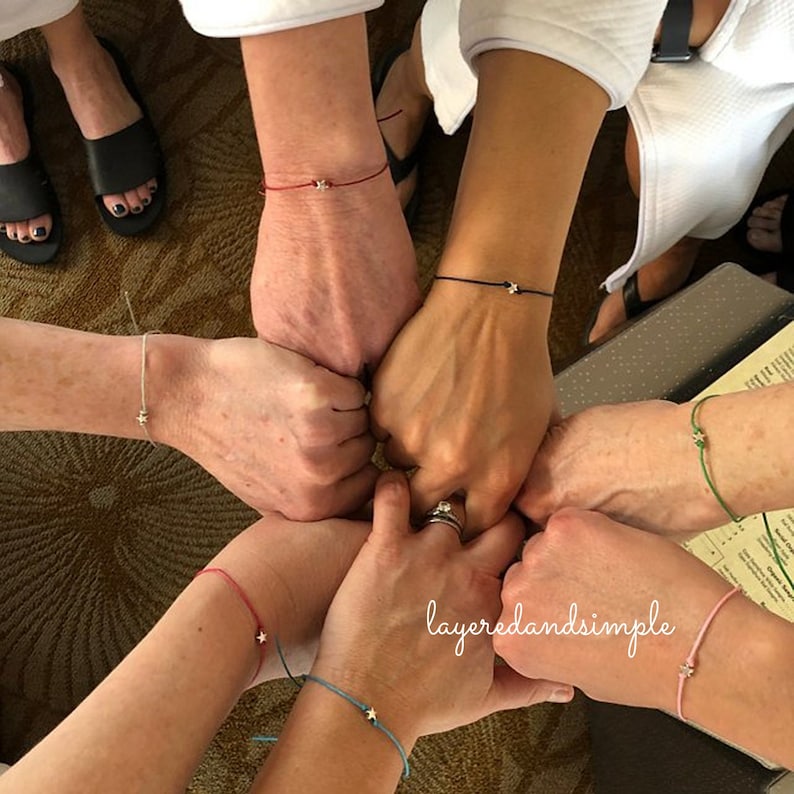 May include: Five wrists with different coloured string bracelets with a small silver star charm on each. The bracelets are layered on top of each other and the hands are clasped together.