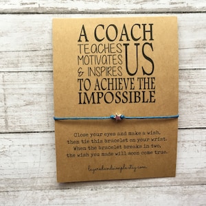May include: A brown card with a blue string bracelet attached. The card reads "A Coach Teaches Motivates & Inspires Us To Achieve The Impossible". The bracelet has a silver star charm with a pink center.