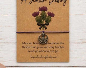 May include: A purple string bracelet with a circular silver-toned metal charm featuring a thistle design. The bracelet is attached to a rectangular kraft paper card printed with the text A Scottish Blessing and a thistle illustration.