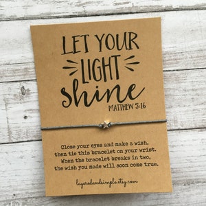 May include: A brown card with the text "LET YOUR LIGHT shine" and "MATTHEW 5:16" printed on it. A gray string bracelet with a silver star charm is tied around the card. The card also includes instructions for making a wish and tying the bracelet.
