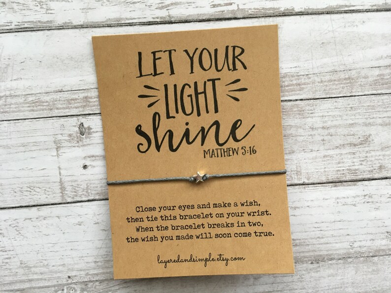 May include: A brown card with the text "LET YOUR LIGHT shine" and "MATTHEW 5:16" printed on it. A gray string bracelet with a silver star charm is tied around the card. The card also includes instructions for making a wish and tying the bracelet.