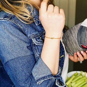 May include: A close-up of a person's wrist wearing a thin orange string bracelet with a small silver charm. The bracelet is on the left wrist of the person wearing a blue denim jacket.